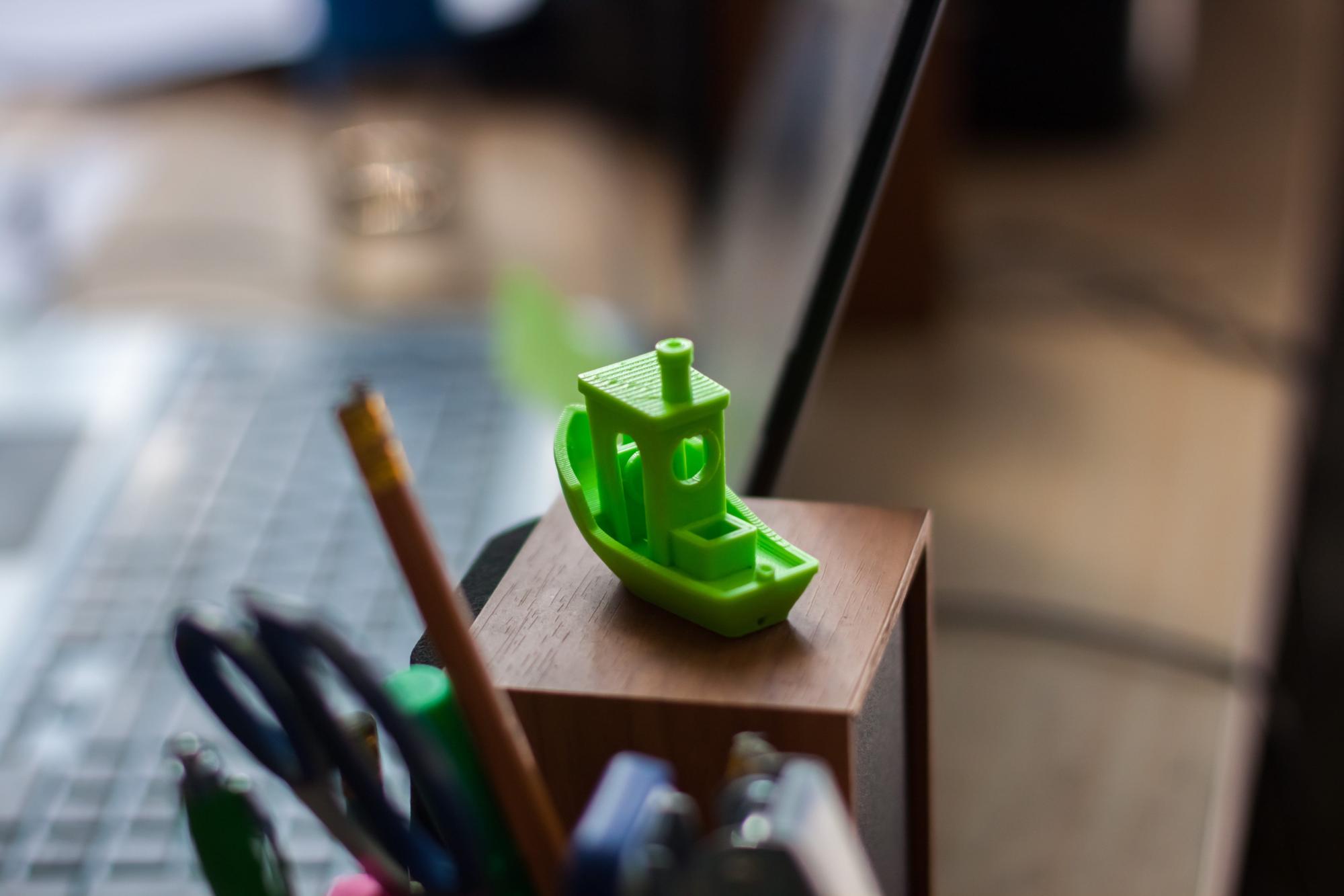 A small 3D printed toy boat on a wooden surface, with office supplies in the foreground.